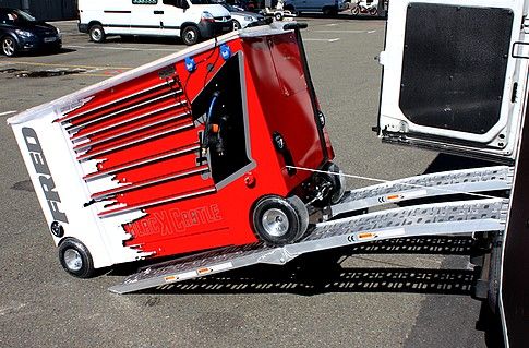 Rode en witte aanhangwagen met gereedschapskist en open laadklep op parkeerplaats, naast bestelwagen.